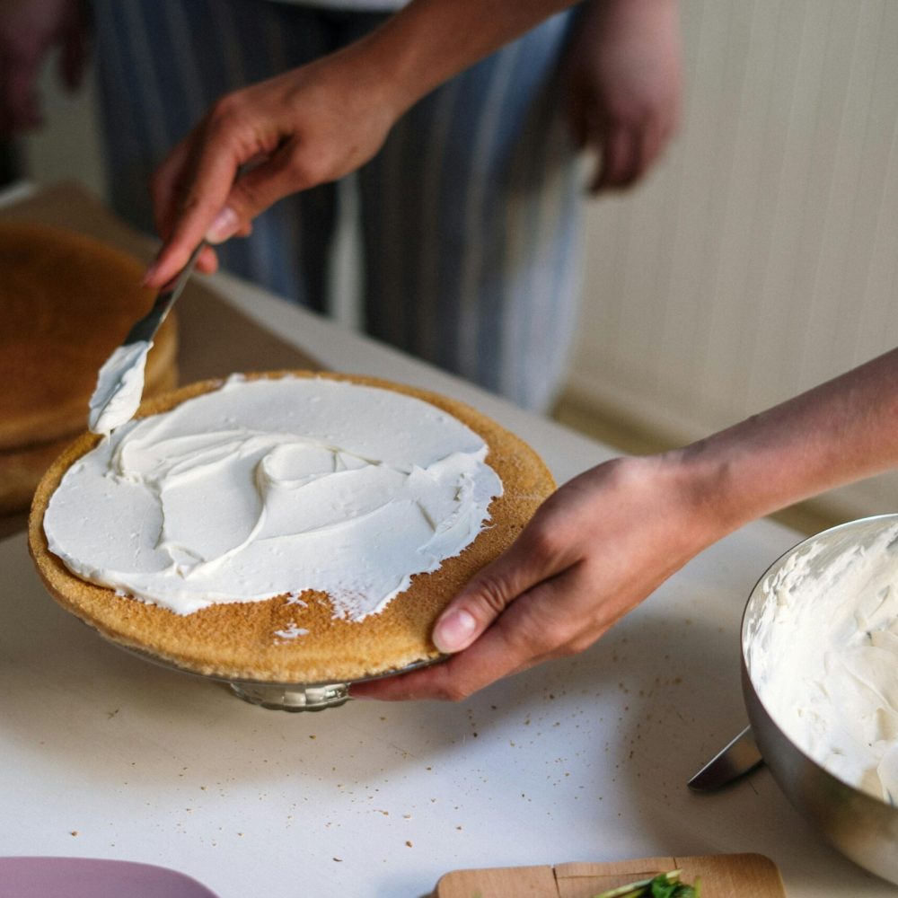 Hands preparing a cake with cream frosting in a cozy kitchen setting, featuring strawberries and a mixing bowl.